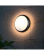 Sfeerfoto van Reality Ambato zwarte ronde wandlamp voor buiten van 27cm in diameter met een wit kunststof schaal aan een muur van betonblokken