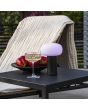 Sfeerfoto van Konstsmide Antibes zwart aluminium tafellamp voor buiten met wit glazen kap van 25 cm hoog op een zwarte tuintafel naast een ligstoel