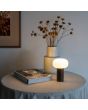Sfeerfoto van Konstsmide Antibes roestbruine aluminium tafellamp voor buiten met wit glazen kap van 25 cm hoog op een bijzettafeltje met een vaas en wat tijdschriften