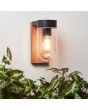 Brilliant Cabar 96372/76 sfeerfoto van wandlamp voor buiten met een houten wandplaat en zwarte bovenzijde en helder glazen kap voor om de lichtbron aan een gestucte buitenmuur