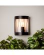Brilliant Reed 44681/63 sfeerfoto van zwarte staande buiten wandlamp met cilindervormig glas en tralies aan spachtelputz muur boven een struik