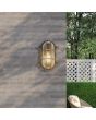 Searchlight Bulkhead 61402BG sfeerfoto van zwartgouden ovale buitenlamp met rooster over ribbelglas aan een grijze muur in een tuin met bomen met wit hekje