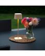 Sfeerfoto van Konstsmide Capri zand kleurige tafellamp op batterijen van 37 cm hoog en touch dimmer op de kop op een zwarte tuintafel  op een rieten onderzetter naast een glazen vaasje met bloemen