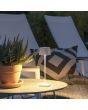 Sfeerfoto van Konstsmide Capri witte oplaadbare aluminium tafellamp voor buiten van 25 cm hoog met een vierkant kapje met touch dimmer op een tuintafel naast twee ligstoelen