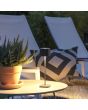Sfeerfoto van Konstsmide Capri zwarte oplaadbare aluminium tafellamp voor buiten van 25 cm hoog met een vierkant kapje met touch dimmer op een tuintafel naast twee ligstoelen