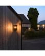Sfeerfoto van Konstsmide Carpi zwarte rechthoekige wandlamp voor buiten van 36,5 cm hoog met transparante ruitjes en een brandend peertje tegen een houten muur van een woning