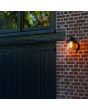 Sfeerfoto van Lutec Cate antiek bruin zwarte wandlamp voor buiten van 22cm hoog met hangende lantaarn van transparant kegelvormig glas naast een staldeur