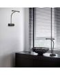 Trio Curtis 579790132 sfeerfoto van zwarte bureaulamp met brede kap met LED strip aan een flexibele hals op een zwart kastje voor een raam met luxaflex en een wandlamp van de zelfde serie aan de wand