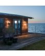 Sfeerfoto van twee Konstsmide Edison mat zwarte wandlamp van 17 cm hoog met cilindervormig staand glas met LED staaf in het midden tegen een houten woning aan zee
