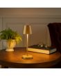 Sfeerfoto van de Brilliant Elora mat goud kleurige oplaadbare tafellamp voor buiten met kunststof bovenzijde voor licht naar boven en beneden met een hoogte van 37 cm en een diameter van 11 cm op een houten bijzettafel