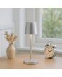 Sfeerfoto van de Brilliant Elora zilver kleurige oplaadbare tafellamp voor buiten met kunststof bovenzijde voor licht naar boven en beneden met een hoogte van 37 cm en een diameter van 11 cm op een houten tafeltje voor een raam