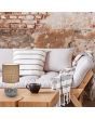 Trio Esna R50651025 sfeerfoto van tafellamp met beige voet met de tekst "home" en een bruine kap van textiel o peen houten salontafel met een landelijke bank met beige kussens voor een ruw stenen muur