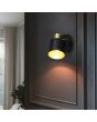 Trio Eva 868870180 sfeerfoto van wandspot met ronde zwarte wandplaat met schakelaar en spotje met goudkleurige fitting en zwart kapje met goudkleurige binnenzijde aan een donkere wand voor een slaapkamer
