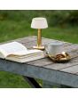 Sfeerfoto van Konstsmide Grasse geborsteld messing oplaadbare tafellamp voor buiten van 20 cm hoog met kapje van textiel op een houten tuintafel naast een boek