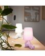Sfeerfoto van lila en witte Reality Huma tafellamp van 24,5 cm hoog met lampvoet in de vorm van een zittend lichaam en een kapje van textiel als hoofd op een houten bureau achter een groene plant