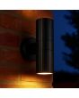 sfeerfoto van een zwart geribbelde up-down wandlamp Keola voor buiten van 18cm hoog met naar beneden en boven gerichte gu10 spotjes tegen een muur in het donker