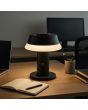 Sfeerfoto van Brilliant Kinich zwarte oplaadbare tafellamp van 16,5 cm hoog en 15 cm in diameter met LED acryl rand onder de kap op een bureau naast een laptop