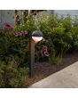 Sfeerfoto van Lutec Looma antraciet grijze sokkellamp voor buiten van 70 cm hoog met een bolvormige kap van 15 cm in diameter met wit kunststof onderzijde in een border met planten