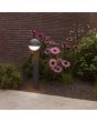 Sfeerfoto van Lutec Looma antraciet grijze sokkellamp voor buiten van 70 cm hoog met een bolvormige kap van 15 cm in diameter met wit kunststof onderzijde in een border met planten langs een pad
