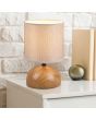 Trio Luci R50351035 sfeerfoto van tafellampje met houtkleurige bolvormige lampvoet van keramiek en beige cilindervormig kapje van textiel op een houten kast met boeken voor een wit stenen muur
