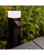 Sfeerfoto van Lutec Mains zwart tuinstopcontact paaltje van 37 cm hoog met twee contactdozen en een brandend LED lichtpunt bovenop in een border met stekker aangesloten