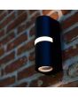 Sfeerfoto van Lutec Mika zwarte wandlamp voor buiten van 19 cm hoog met up en down light en LED rand halverwege de lamp tegen een rood ruw stenen muur