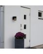 Sfeerfoto van Konstsmide Pistoia zwarte wandlamp voor buiten van 17,5 cm hoog met een buisvormig kapje met streepvormige openingen voor het licht naast een voordeur