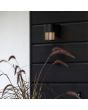 Sfeerfoto van Konstsmide Pistoia zwarte wandlamp voor buiten van 17,5 cm hoog met een buisvormig kapje met streepvormige openingen voor het licht tegen een houten schutting boven een plant