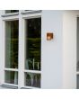 Sfeerfoto van Konstsmide Pistoia roestbruine wandlamp voor buiten van 17,5 cm hoog met een buisvormig kapje met streepvormige openingen voor het licht tegen een gevel naast een raampartij