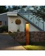 Sfeerfoto van een Konststmide Rho roestbruine rechthoekige wandlamp van 28 cm hoog met geintegreerde LED met lichtbundel langs een metalen plaat tegen de gevel van een bungalow met een paallamp op de voorgrond uit dezelfde serie