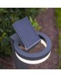 Sfeerfoto met bovenaanzicht van Lutec Runda antraciet grijze sokkellamp van 74 cm hoog met schijfvormige lantaarn met opgeklapt kantelbaar zonnepaneel bovenop in een tuin
