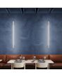 Sfeerfoto van twee smalle beige wandlampen Sappo van 150 cm lang met LED voor indirect licht tegen een blauwe muur in een restaurant