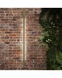 Sfeerfoto van een smalle olijfgroene wandlamp Sappo van 120 cm lang met LED voor indirect licht tegen een rood stenen muur naast beplanting