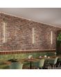 Sfeerfoto van drie smalle olijfgroene wandlampen Sappo van 150 cm lang met LED voor indirect licht tegen een rood stenen muur in een restaurant
