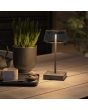 Sfeerfoto van Konstsmide Scilla zwarte tafellamp voor buiten op batterijen met transparant glazen kap met touch dimmer op een tuintafel met groene plant in een pot