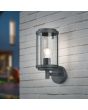 Trio Tanaro 202360142 sfeerfoto van antraciet grijze wandlamp voor buiten met cilindervormige staande kap met vier dubbele spijlen en helder glas aan een wit stenen muur