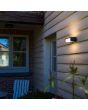 Sfeerfoto van Lutec Telis antraciet grijze wandlamp voor buiten van 18 cm breed met zonnepaneel bovenop en wit kunststof venster naar beneden en voren en klein venstertje naar boven tegen een houten wand van een woning